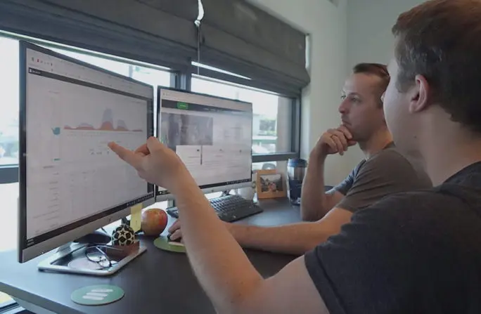 Two men analyzing data on dual monitors in a modern office setting, focusing on graphs and charts.