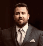 Man in a suit with a pink tie and pocket square against a dark background.