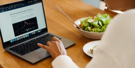 A woman's hand rests on the mousepad of a laptop. Next to the laptop is a bowl of salad.