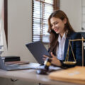A young female lawyer sits at a desk, holding a tablet. On her desk is an open laptop, a gavel, justic scales, and an orange legal book