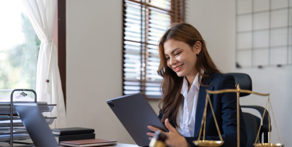 A young female lawyer sits at a desk, holding a tablet. On her desk is an open laptop, a gavel, justic scales, and an orange legal book