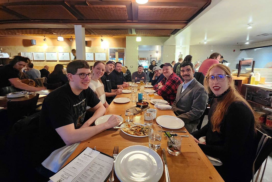 Group of people dining at a long wooden table in a cozy restaurant setting, enjoying food and drinks.