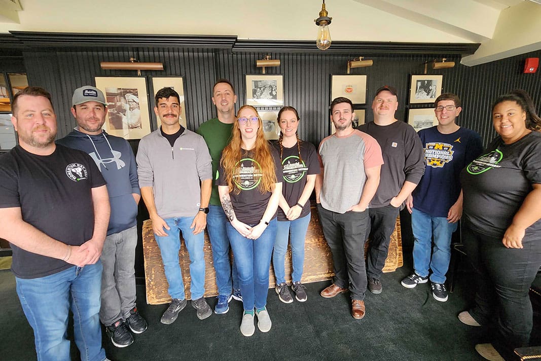 Group of ten people smiling indoors, standing in a line against a dark wall with framed images.